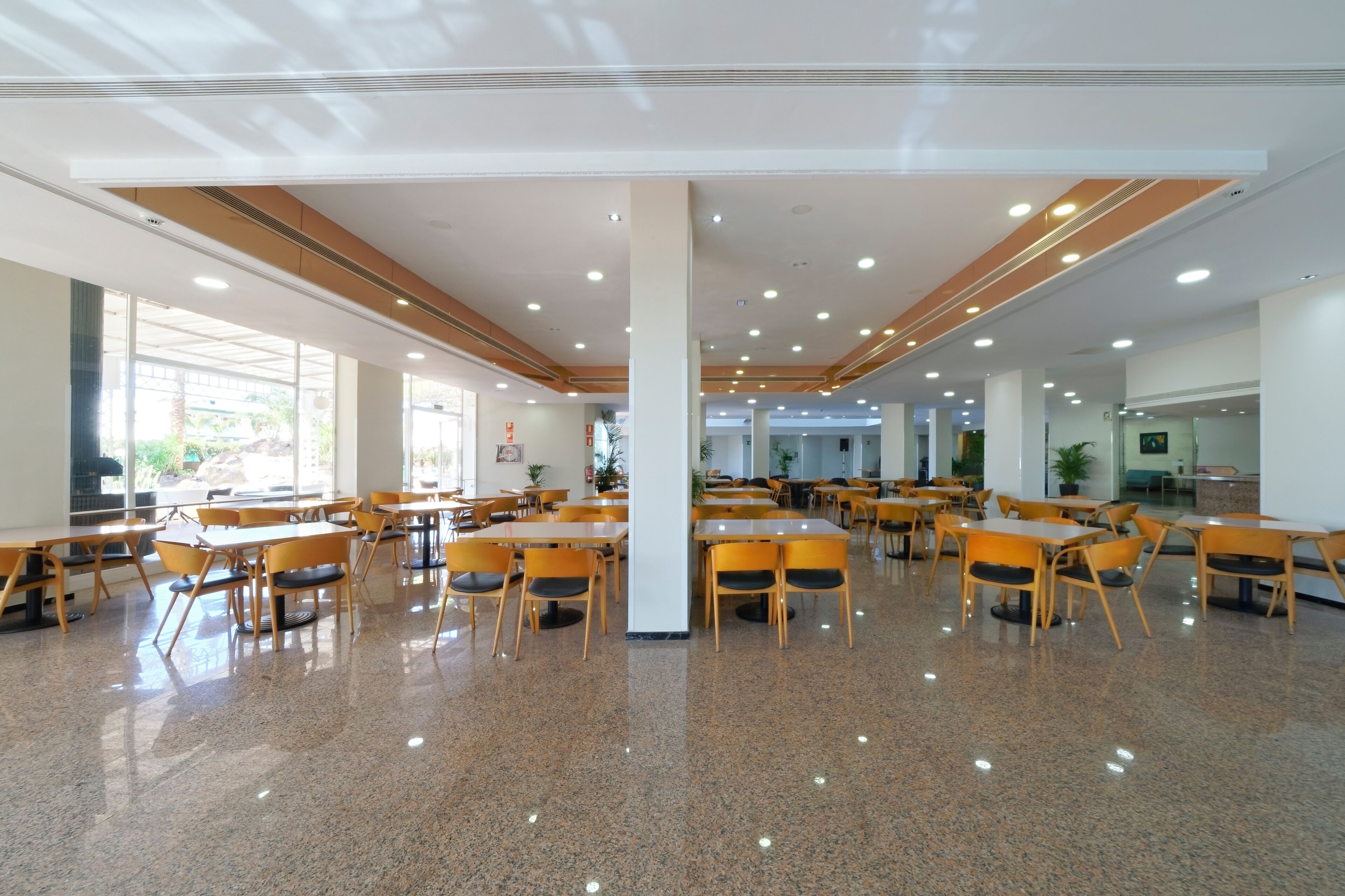 A spacious cafeteria with multiple tables and chairs, large windows, and a polished floor.