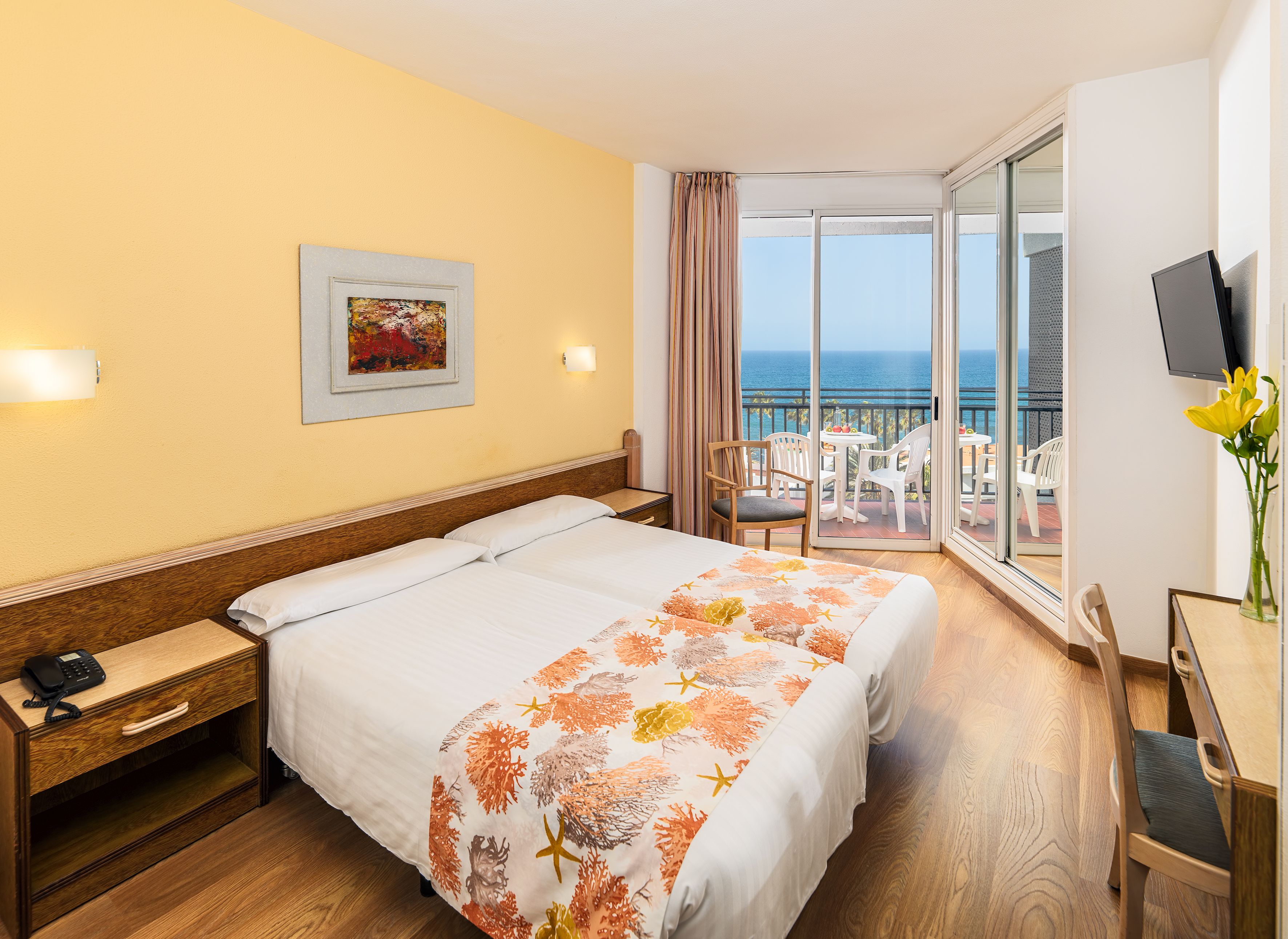 Une chambre d’hôtel avec un lit, un bureau et une chaise, un téléphone, un tableau au mur et un balcon avec vue sur la mer.