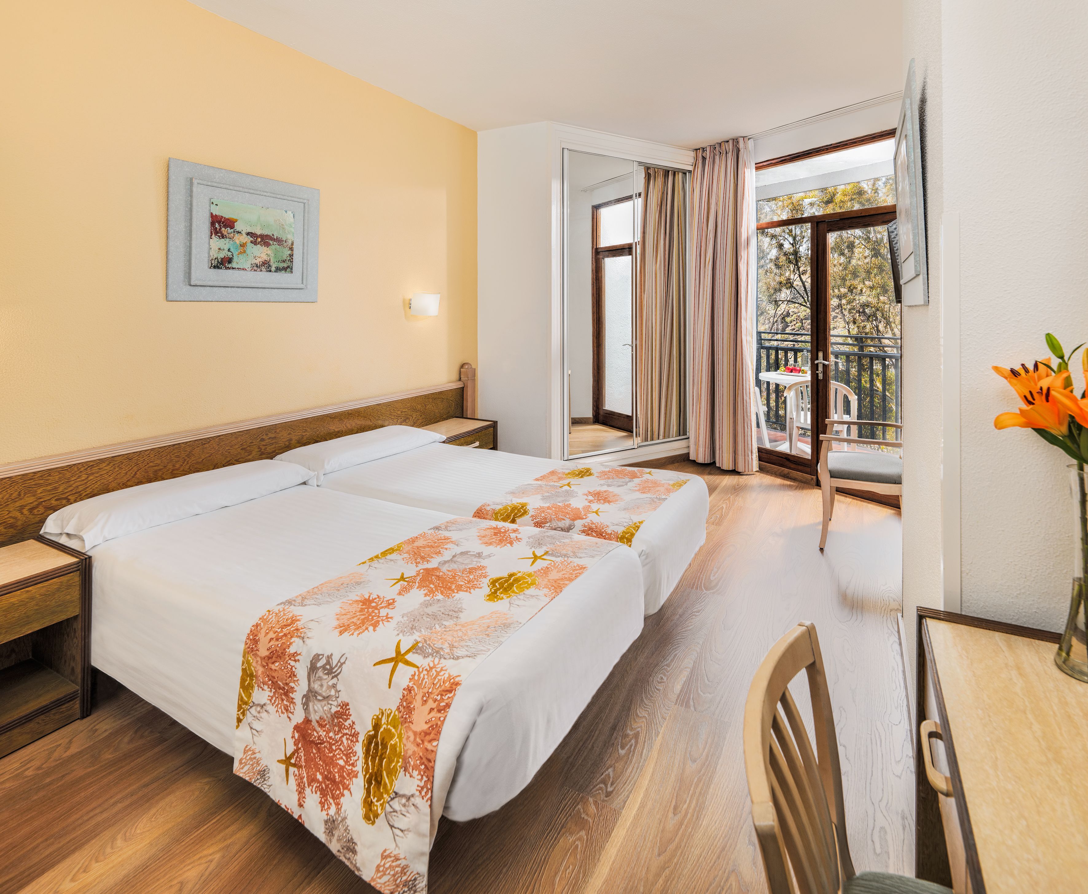A hotel room with two beds, a desk, a chair, a window with curtains, a small table, and a framed picture on the wall.