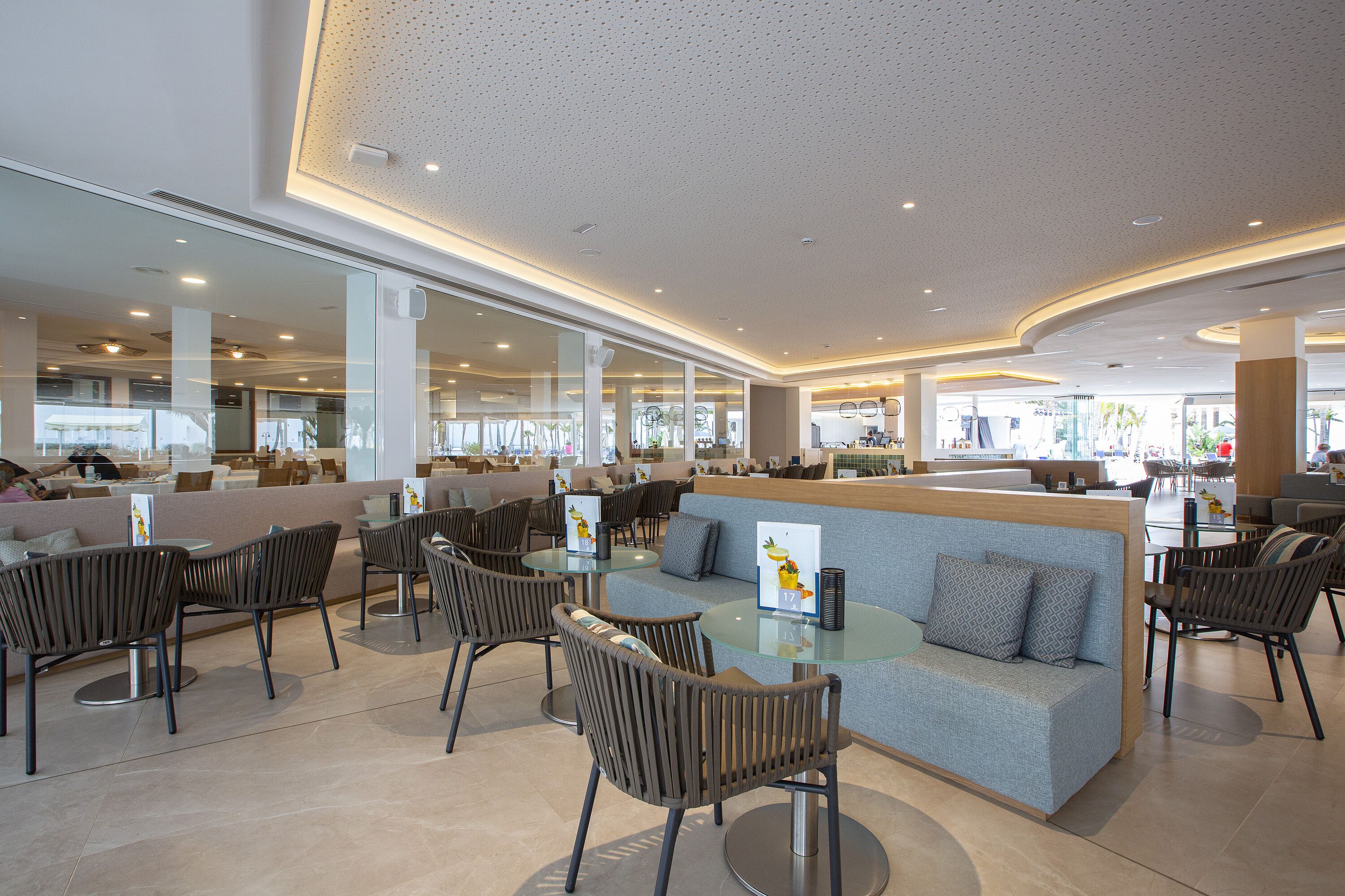 A modern indoor seating area with round tables and cushioned seating, large windows, and a high ceiling with recessed lighting.