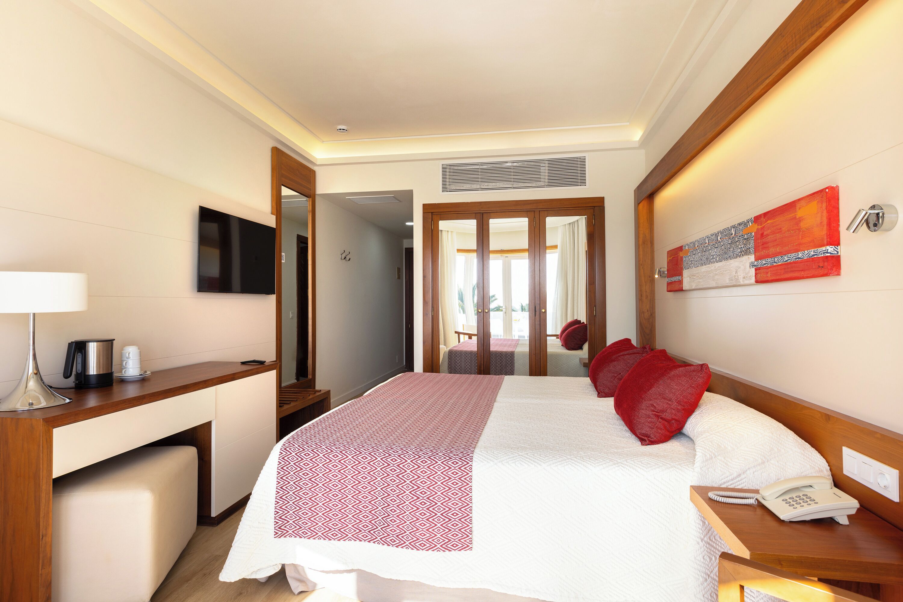Habitación de hotel con cama, mesita de noche con teléfono, escritorio con lámpara, cafetera y televisor en la pared.