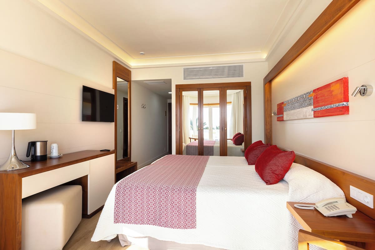 Habitación de hotel con cama, mesita de noche con teléfono, escritorio con lámpara, cafetera y televisor en la pared.