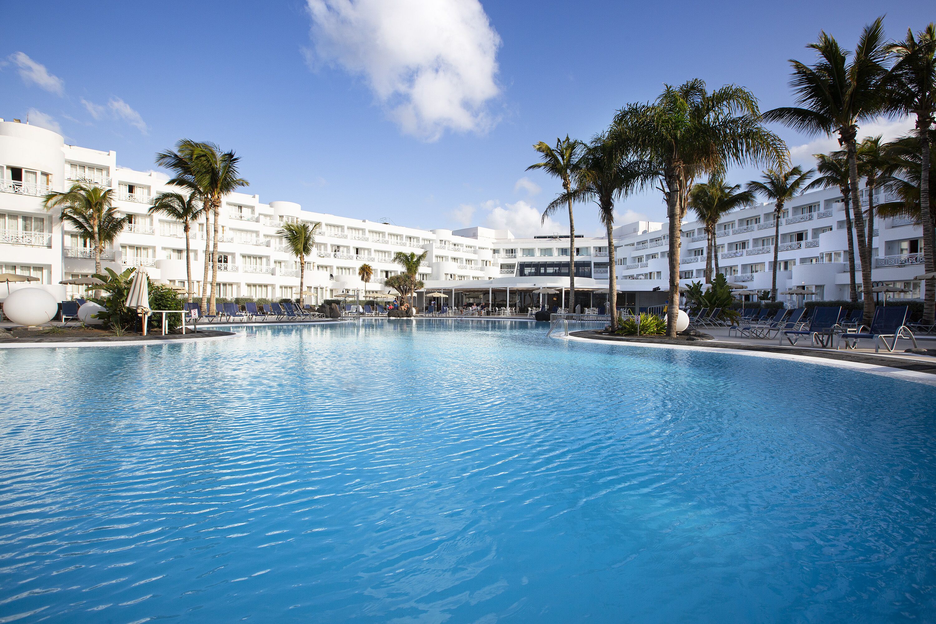 Una gran piscina con sillas de descanso y palmeras, rodeada por un edificio blanco con varios balcones.