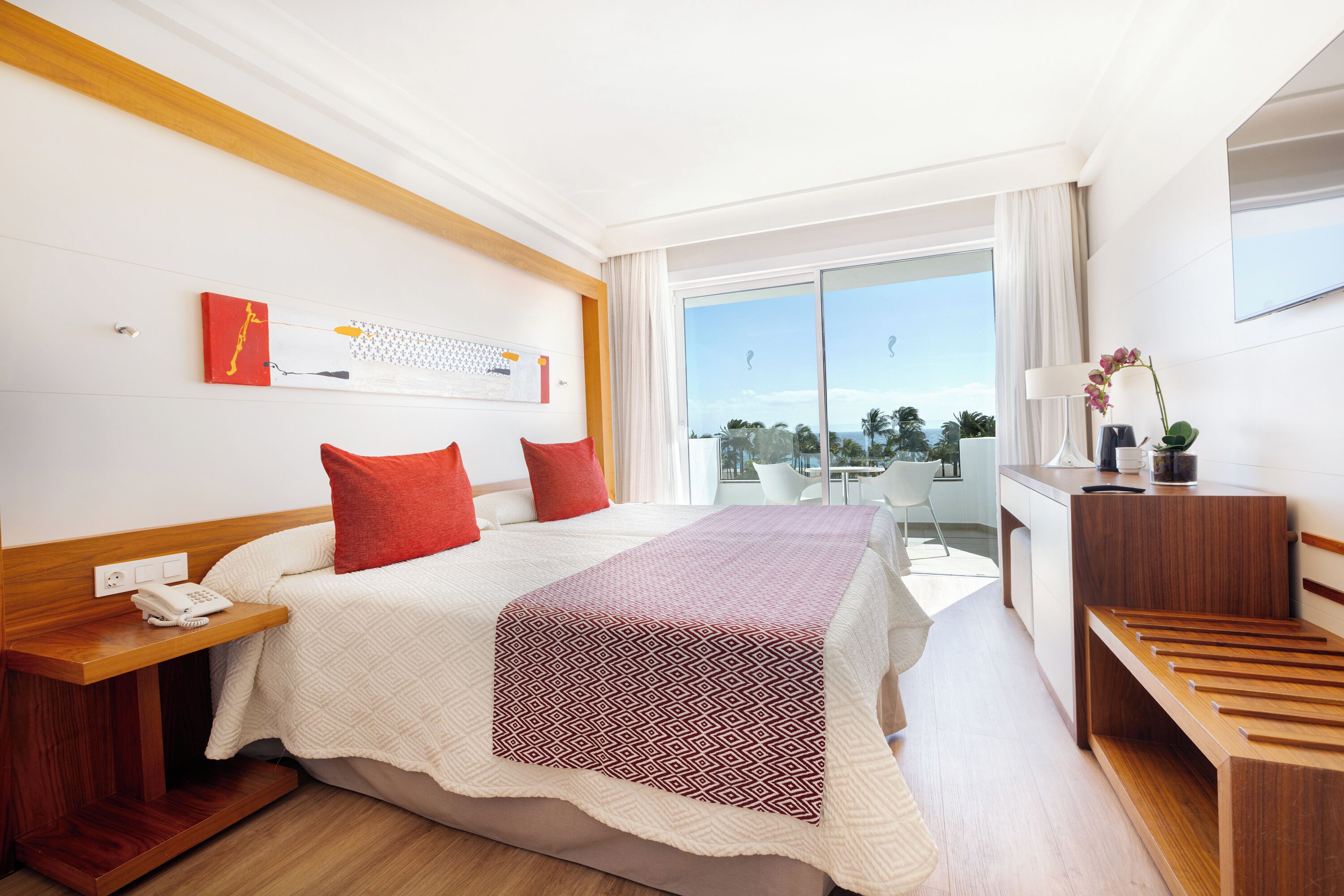 Habitación de hotel con cama, mesita de noche y escritorio con lámpara. Se ve una ventana con vistas a palmeras y un balcón.