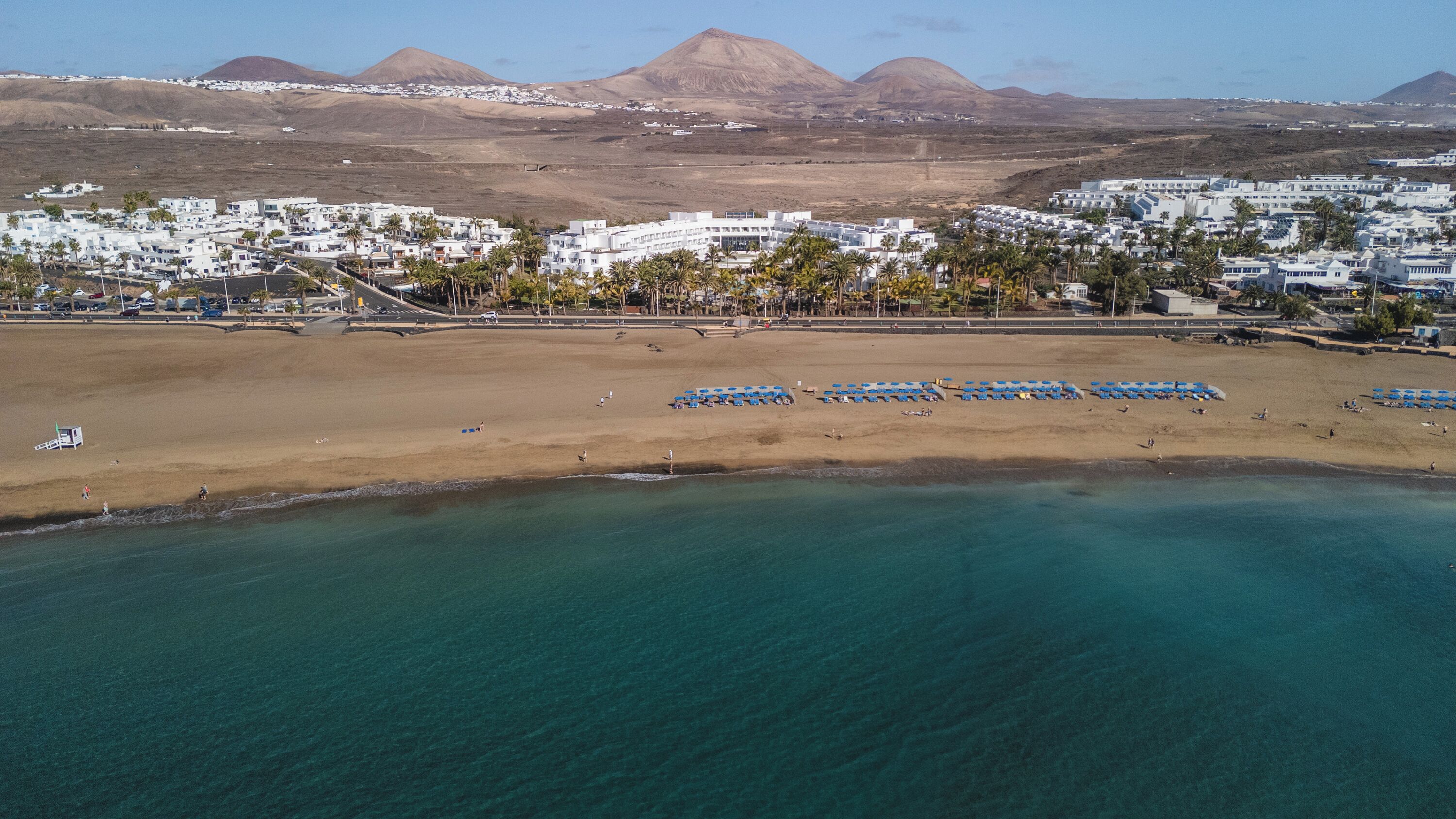 Un pueblo costero con una playa, aguas azules cristalinas y edificios blancos con palmeras.