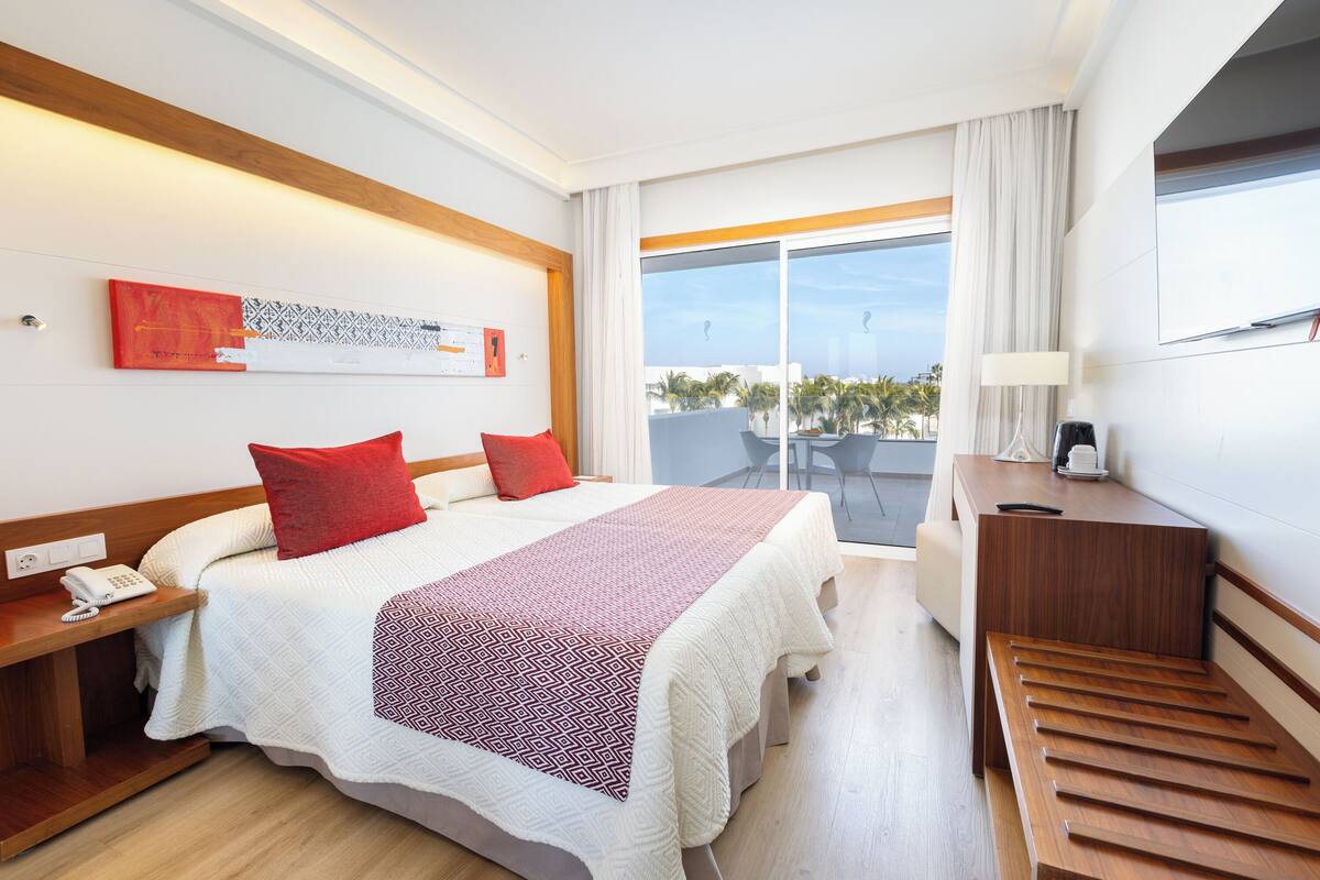 A hotel room with a bed, a desk, a chair, a phone, and a window with a view of palm trees.