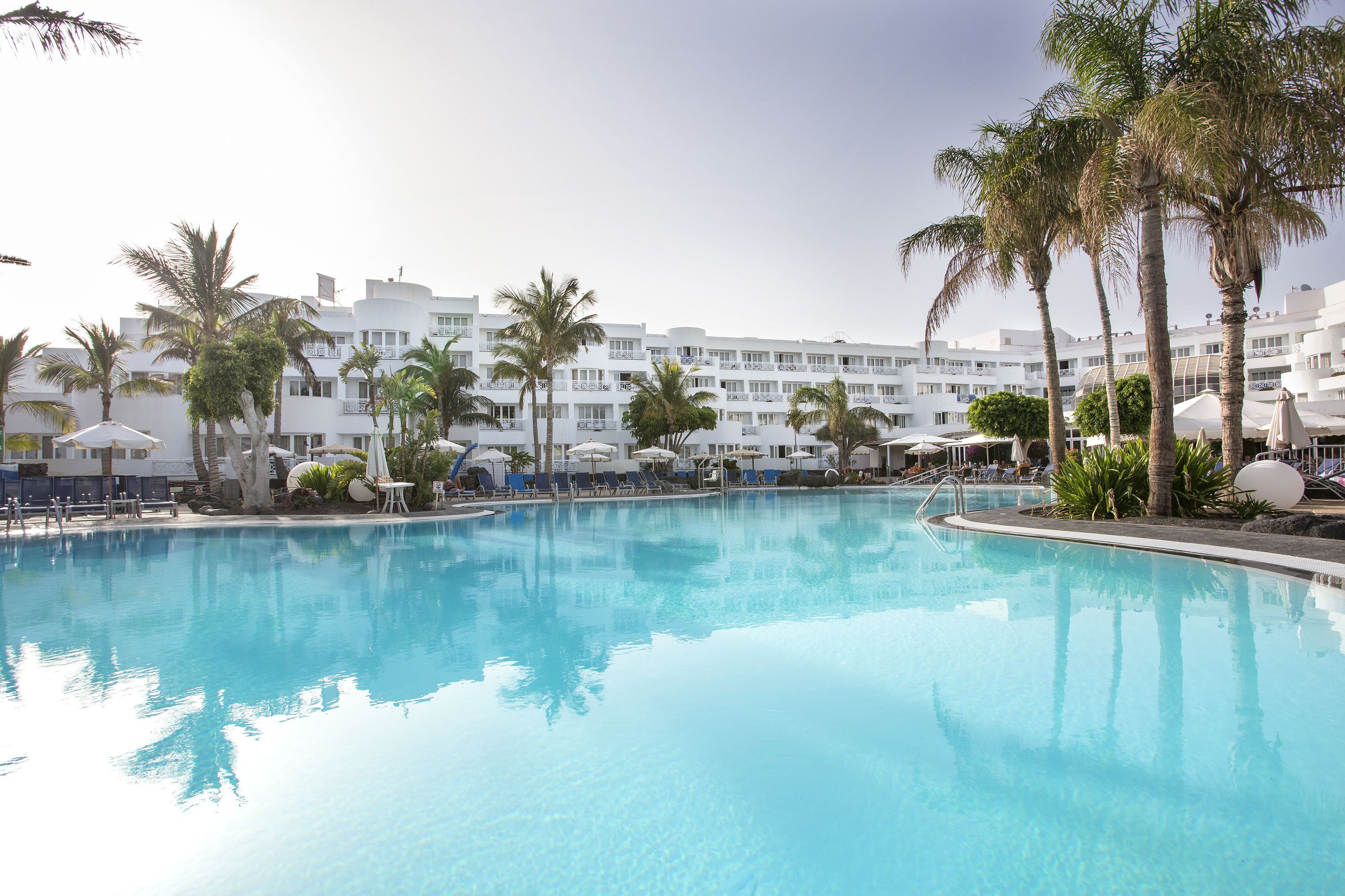 A swimming pool with lounge chairs and umbrellas, surrounded by palm trees and a hotel building in the background.