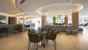 A modern lobby with a round glass table surrounded by wicker chairs, a seating area with sofas, and a reception desk. - Hipotels La Geria (Tías)