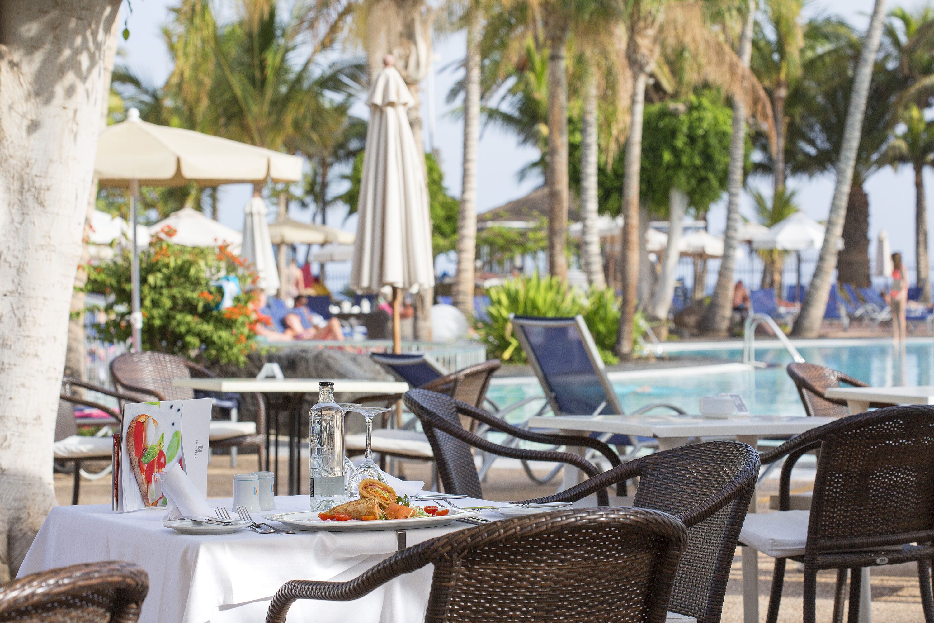 Un área de comedor junto a la piscina con sillas de mimbre, una mesa dispuesta con platos, cubiertos y una botella.