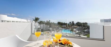 Una mesa de comedor con vista a una piscina y palmeras, una jarra de jugo de naranja y una bandeja de frutas.
