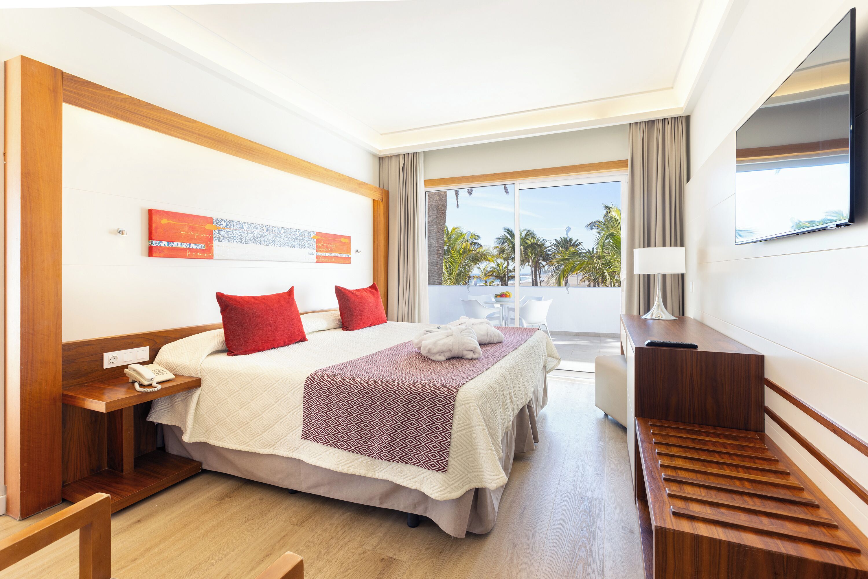 A hotel room with a bed, a desk, a TV, and a balcony with a view of palm trees.