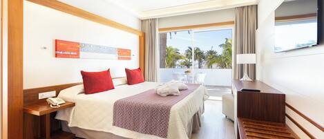A hotel room with a bed, a desk, a TV, and a balcony with a view of palm trees.