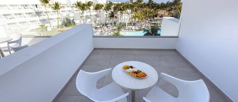 A balcony with a table and chairs, overlooking a pool area with palm trees and a building in the background.