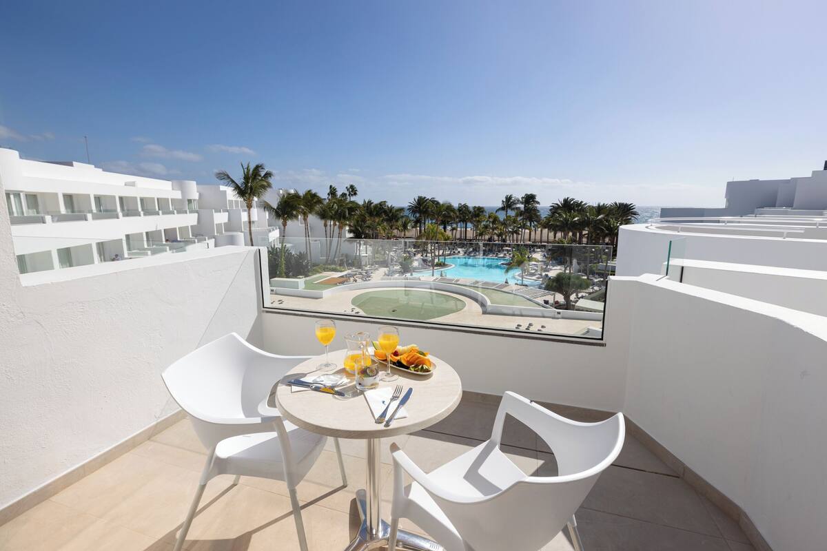 A balcony with a table set for two, overlooking a pool and palm trees.