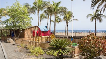 Kids can play in the beachfront playground with a wooden playhouse and slide, surrounded by a fence.