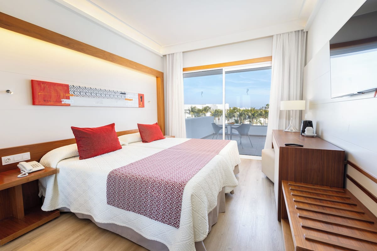 Habitación de hotel con cama, escritorio, silla, teléfono y ventana con vista a palmeras.