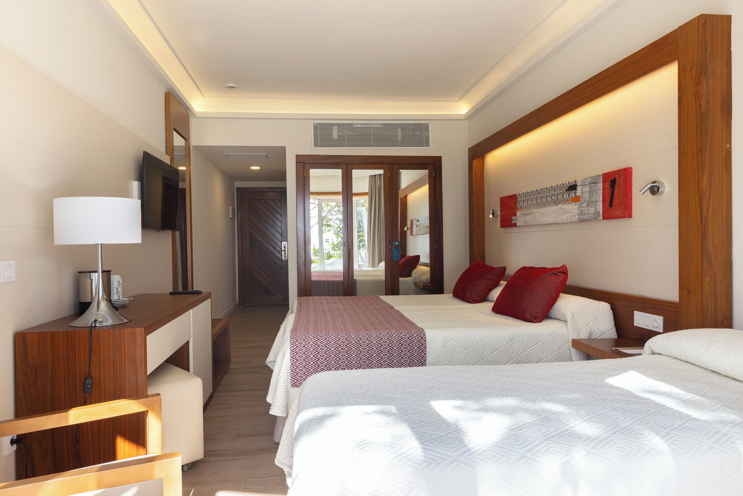 A hotel room with two beds, a desk with a lamp, a TV, and a large mirror.
