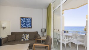 Una sala de estar moderna con sofá, una mesa de centro y un comedor con sillas. Un cuadro adorna la pared, y se divisa una vista al mar desde el balcón.