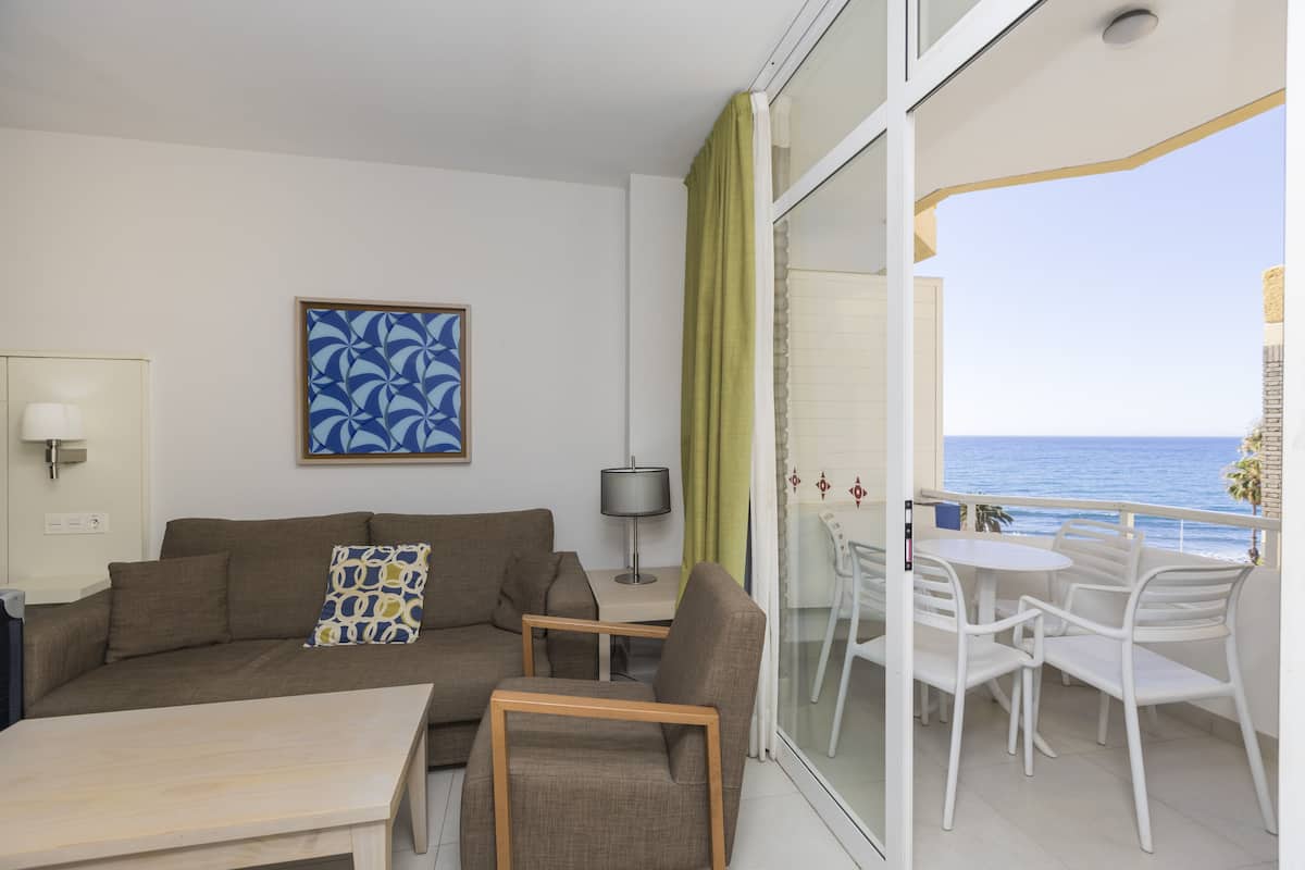 Un salon moderne avec un canapé, une table basse et un coin repas avec des chaises. Un tableau est accroché au mur, et on peut admirer la mer depuis le balcon.