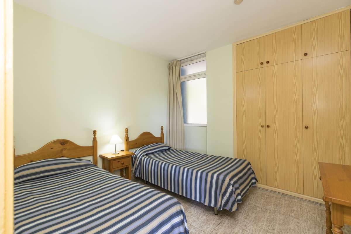 Une chambre avec deux lits simples, une armoire en bois, une petite table et une fenêtre avec des rideaux.