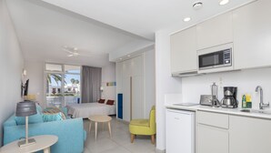 Una habitación de hotel moderna con kitchenette, una cama con almohadas de colores, un pequeño comedor y vistas a palmeras.