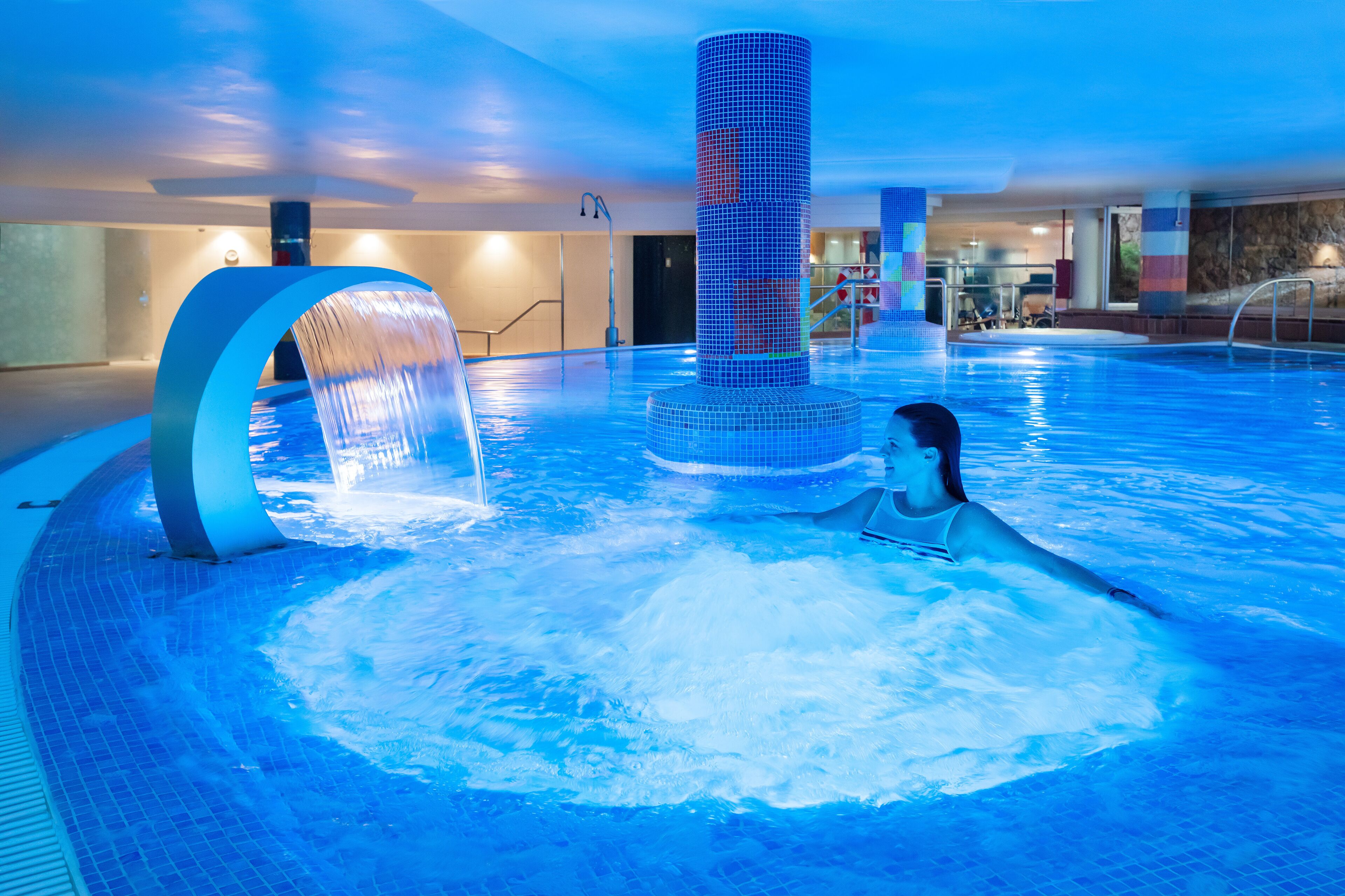 A large indoor swimming pool with a central water feature and a person relaxing in the water.