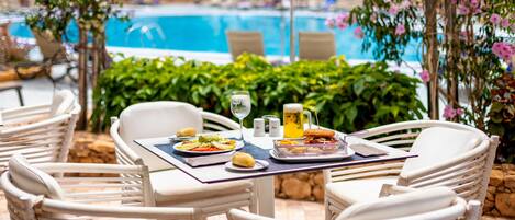 An outdoor dining area with white wicker chairs and a table set for two, overlooking a pool.