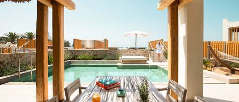 A wooden deck with a pool, lounge chairs, and a table with a drink and a plant.