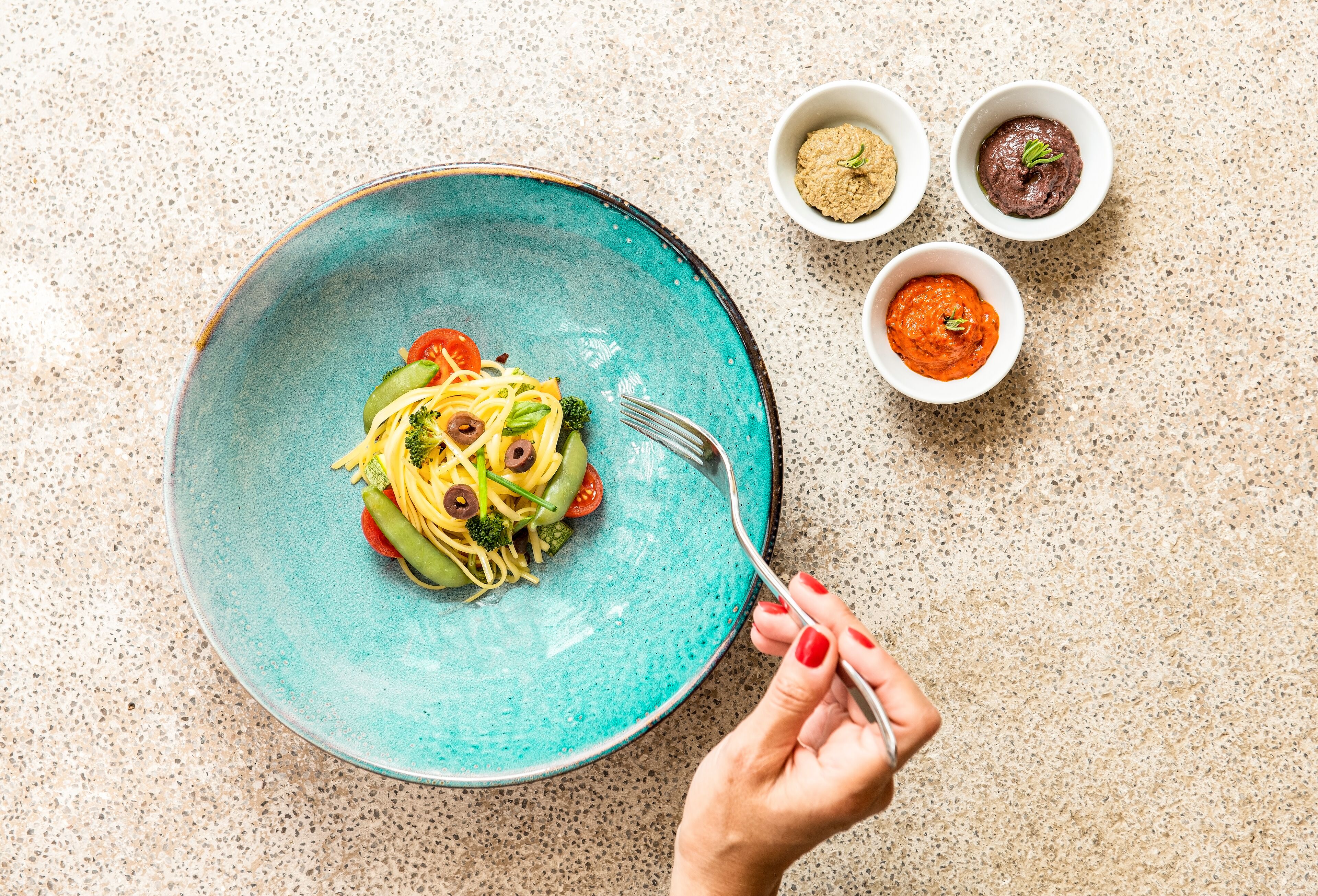 Un plato de pasta con verduras y dos recipientes pequeños de salsa sobre una encimera con motas.