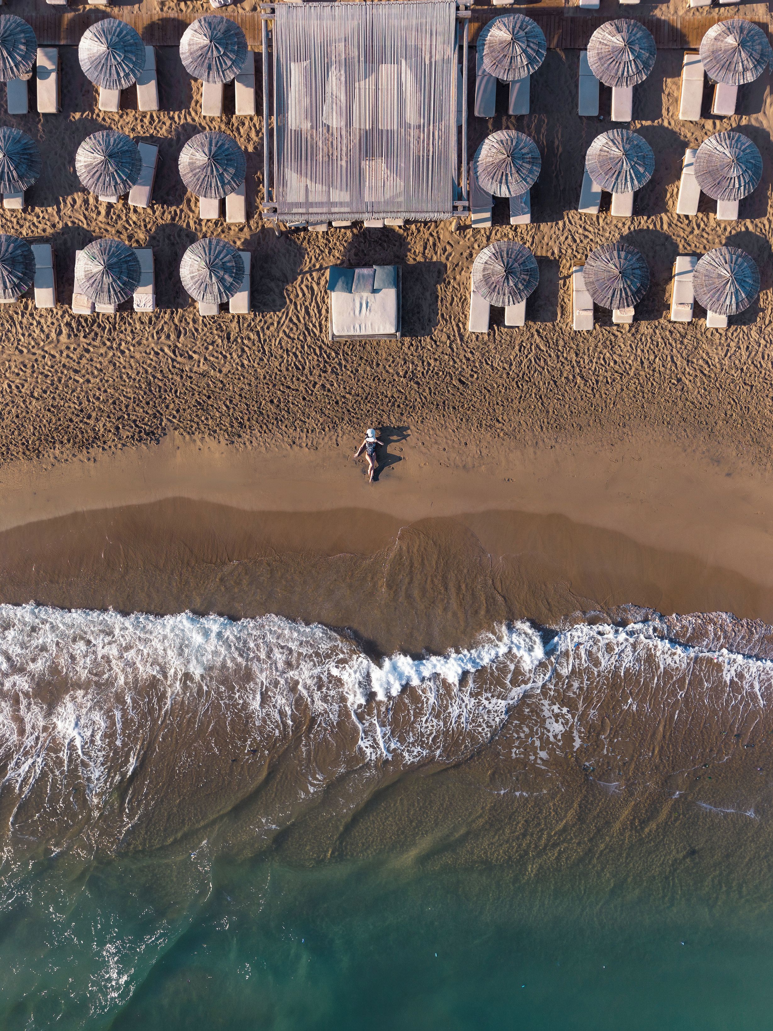 Une vue aérienne d’une plage, avec des rangées de parasols et de chaises longues, une personne qui marche sur le sable et des vagues qui se rapprochent du rivage.