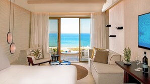 A hotel room with a large bed, a sofa, a dining table, and a TV. There is a view of the ocean through the windows. - Mitsis Selection Rinela (Hersonissos)