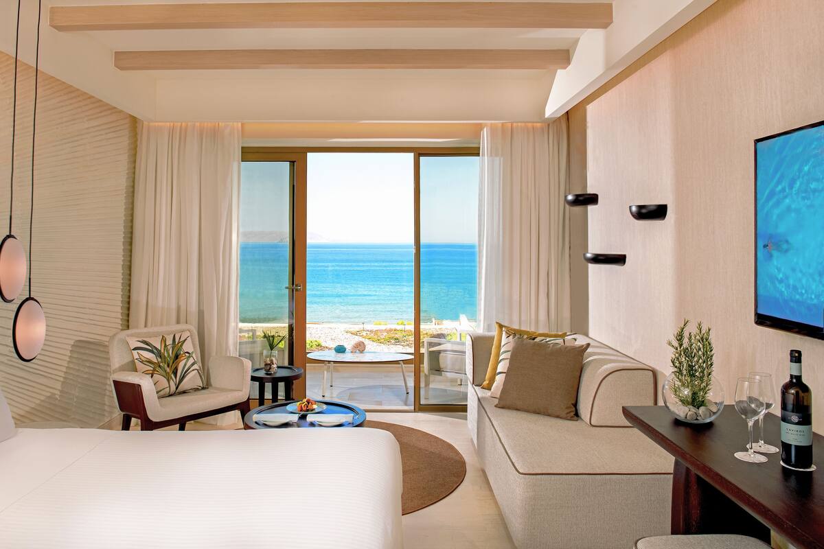 Une chambre d’hôtel avec un grand lit, un canapé, une table à manger et un téléviseur. Les fenêtres offrent une vue sur l’océan.