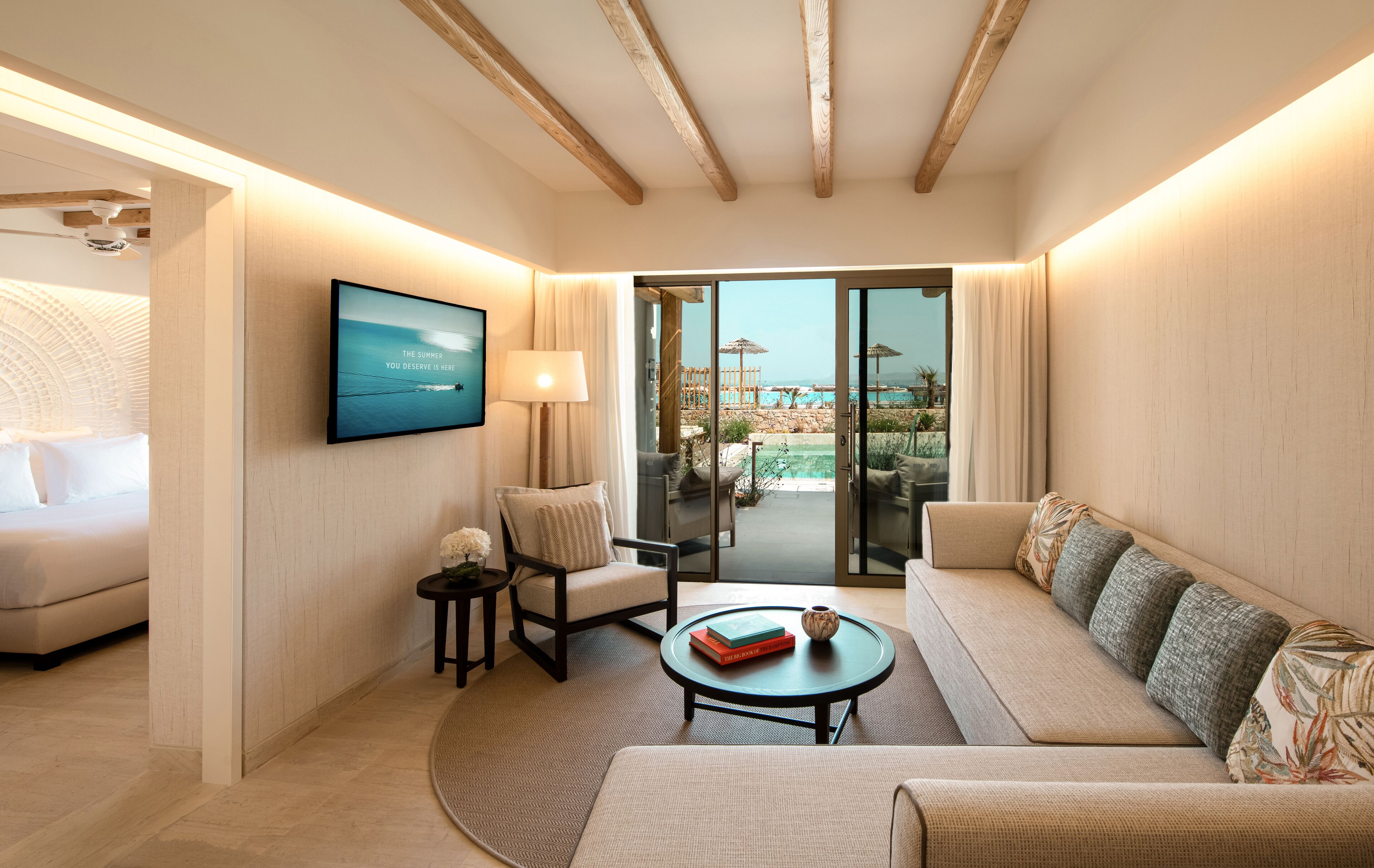 A modern hotel room with a sofa, a chair, a small table with books, a TV on the wall, and a sliding glass door leading to an outdoor area with a pool.
