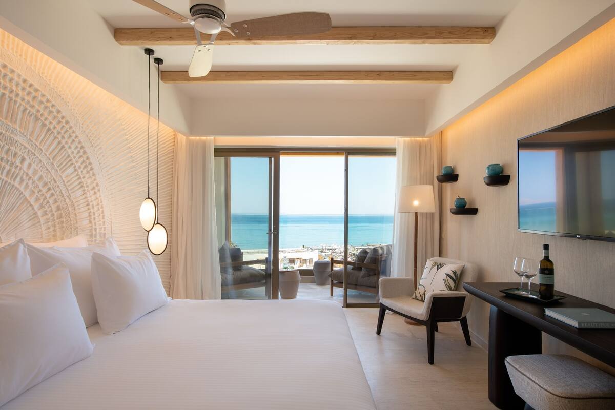 Une chambre d’hôtel avec un grand lit, un ventilateur de plafond, une télévision, un bureau avec une bouteille et des verres, et une vue sur la plage à travers la porte vitrée.