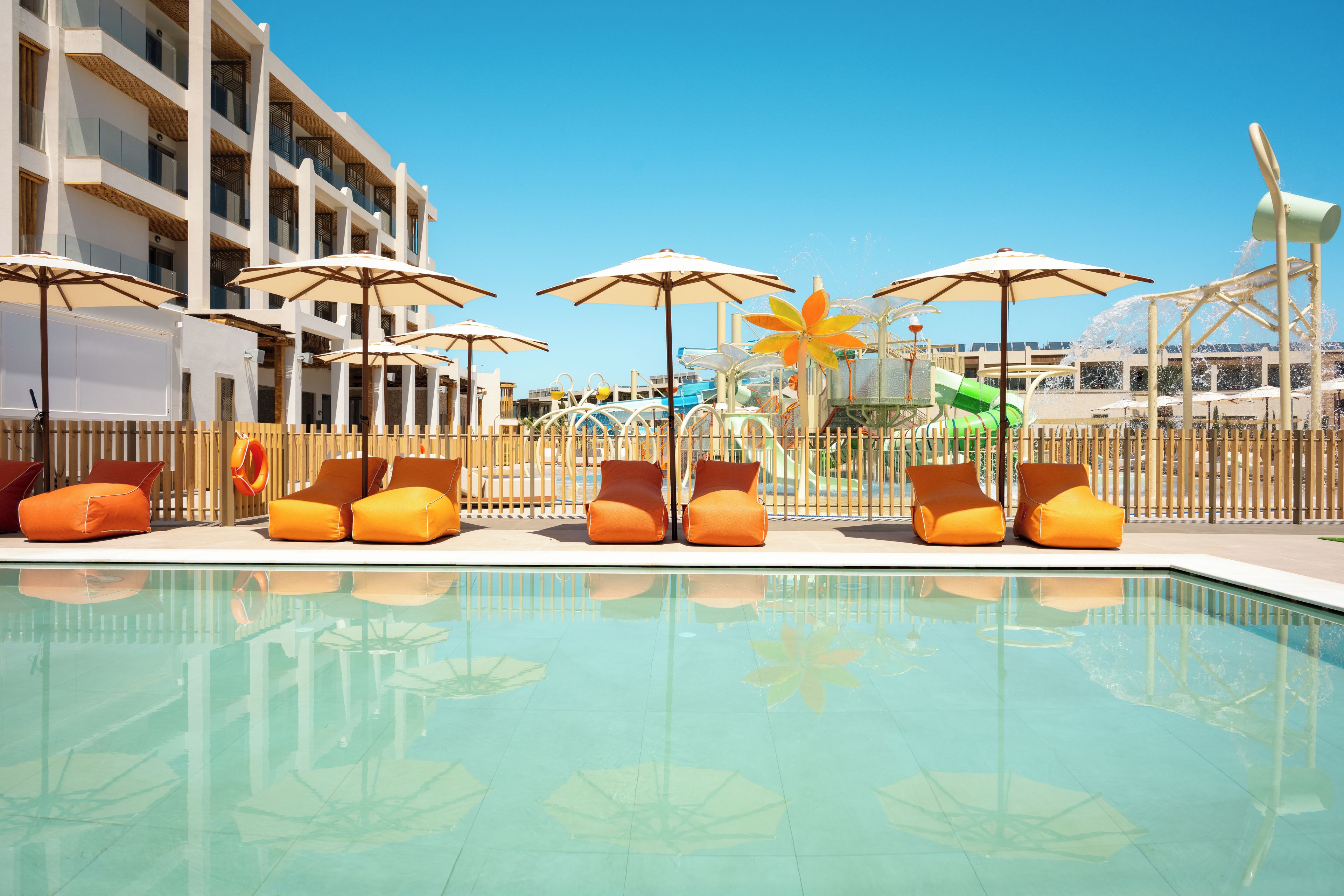 A pool area with lounge chairs and umbrellas, a clear blue sky, and a building in the background.
