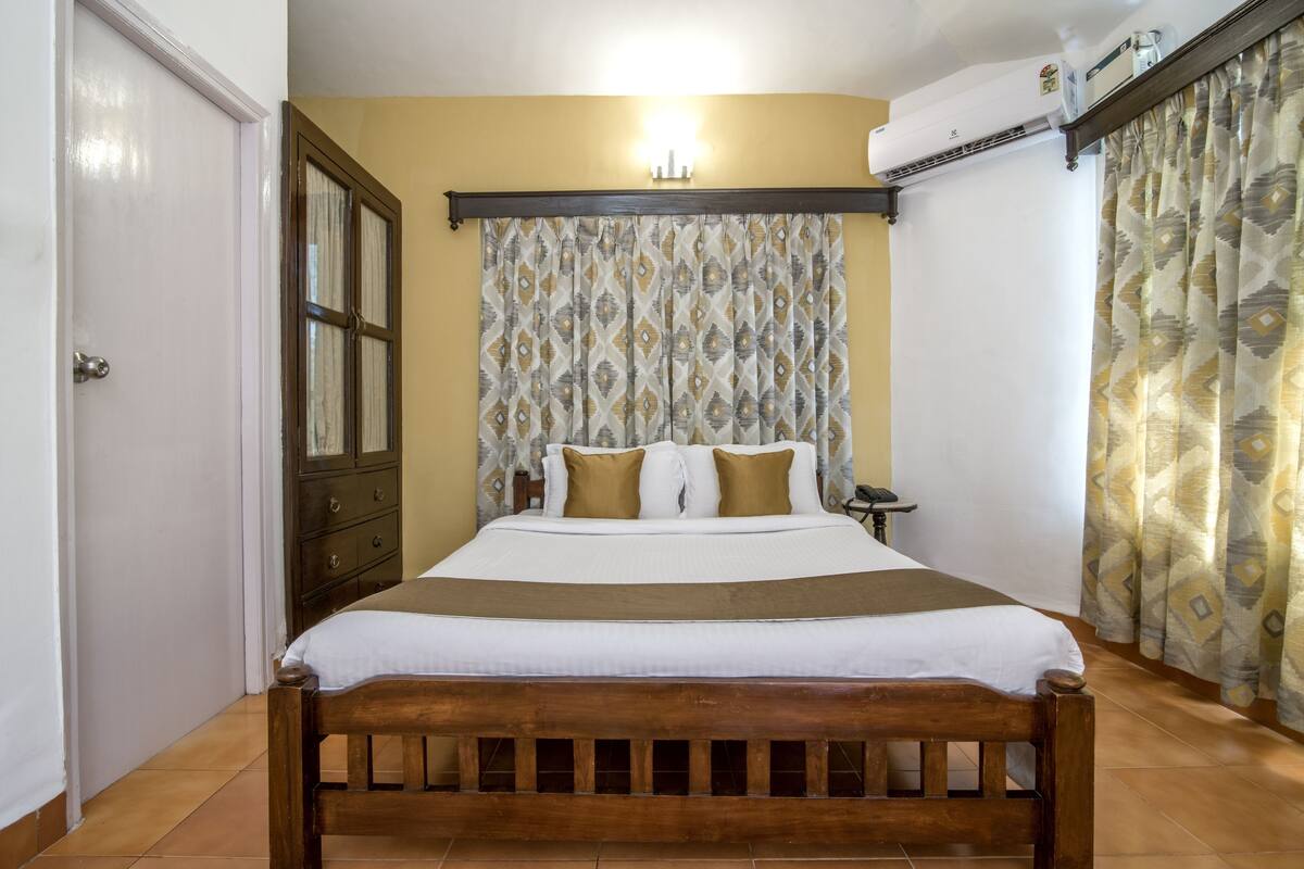 Un dormitorio con cama de madera, una mesita de noche, aire acondicionado de pared y cortinas con estampado.