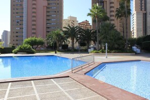 A swimming pool area with a tiled deck, surrounded by palm trees and tall residential buildings. - Vistamar Apartamentos (Benidorm)