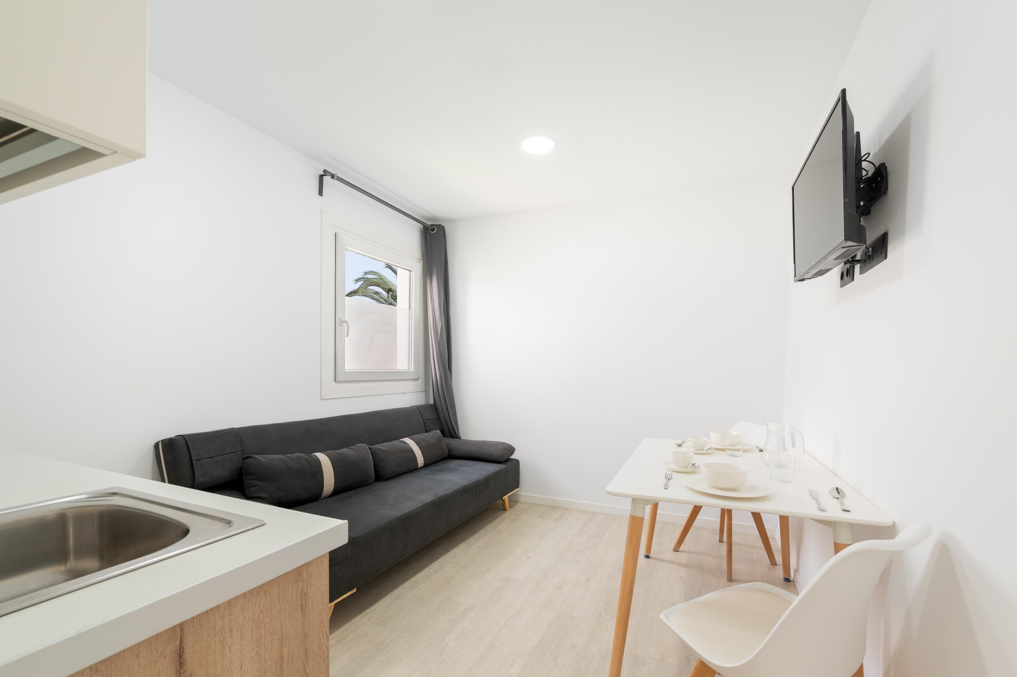 A compact living space with a kitchenette, a sofa, a dining table, and a TV mounted on the wall.