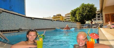 A pool area with two people relaxing, a drink on the side, and a view of buildings in the background.