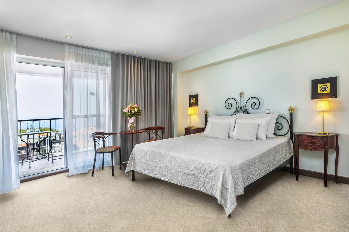 Ein Hotelzimmer mit einem großen Bett, zwei Nachttischen, einem Sessel, einem kleinen Tisch mit Blumenvase und einem Balkon mit Gartenmöbeln.