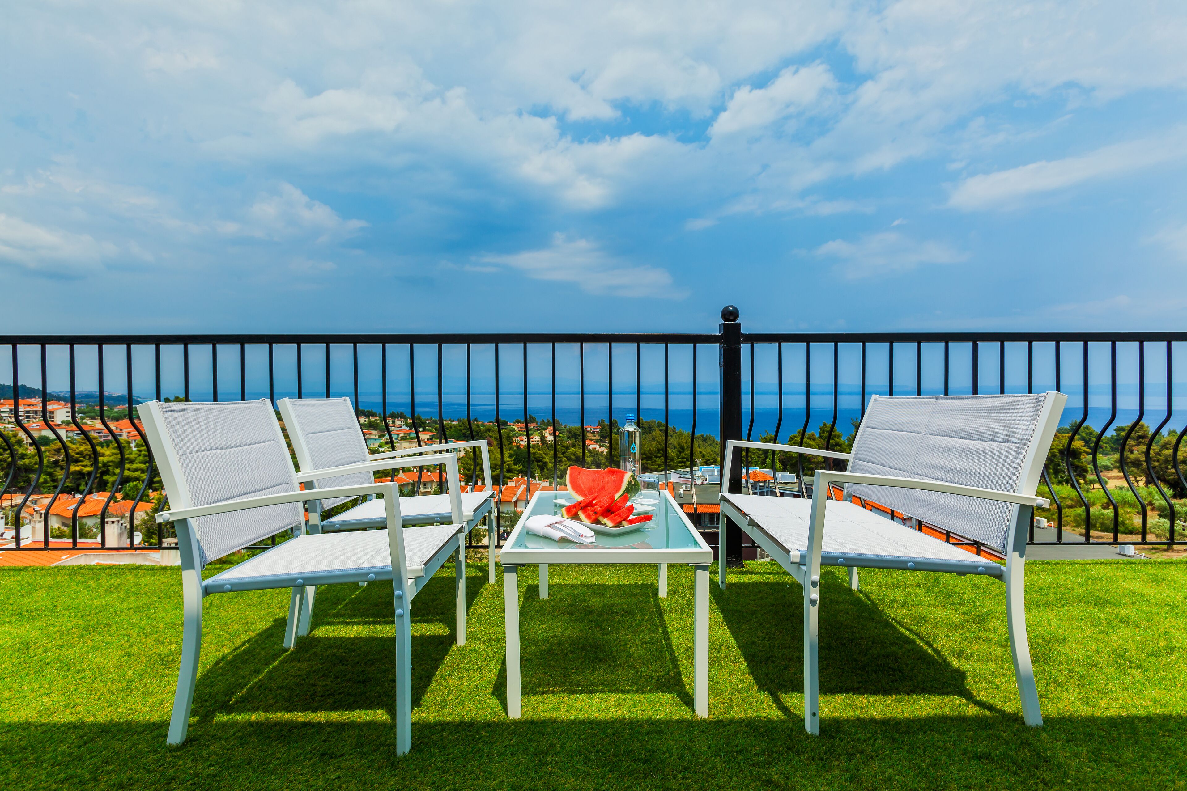 Ein Balkon mit zwei weißen Stühlen und einem Tisch, der Blick auf die Küste mit Häusern im Hintergrund freigibt.