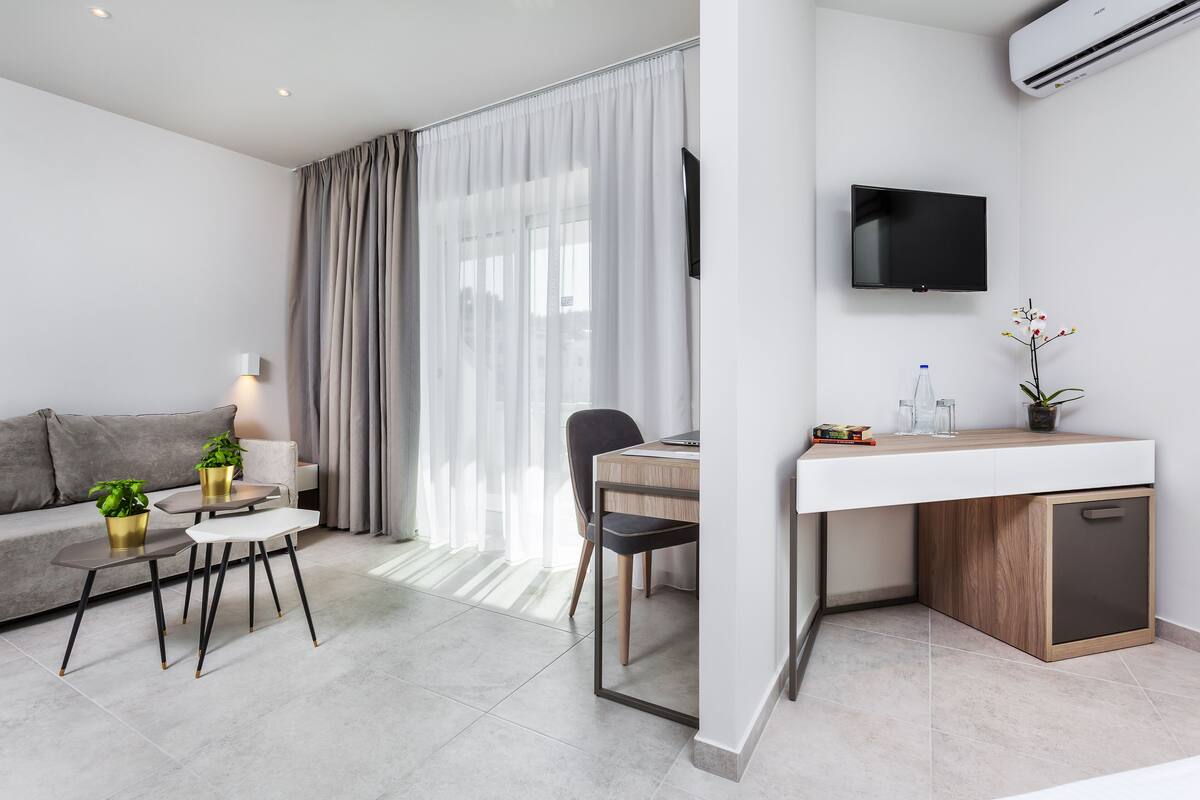 Ein modernes Hotelzimmer mit einer Couch, einem Esstisch mit Stühlen, einer kleinen Küchenzeile und einem an der Wand befestigten Fernseher.