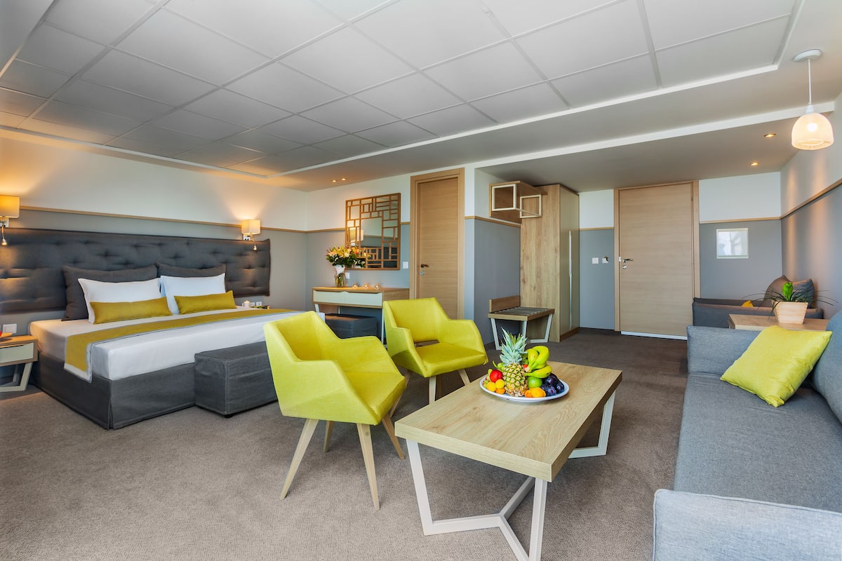 Ein modernes Hotelzimmer mit einem großen Bett, einer Couch, einem Esstisch und leuchtend gelben Stühlen.