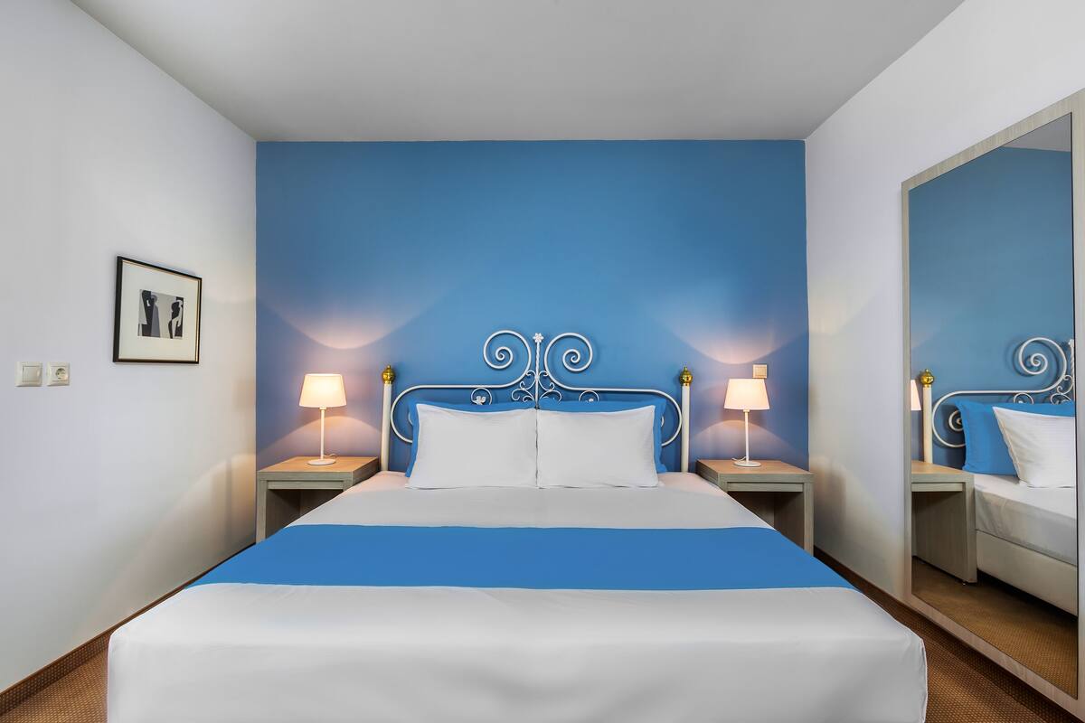 Ein ordentlich bezogenes Bett mit blauer Bettdecke, zwei Nachttischlampen und einem gerahmten Bild an der Wand.