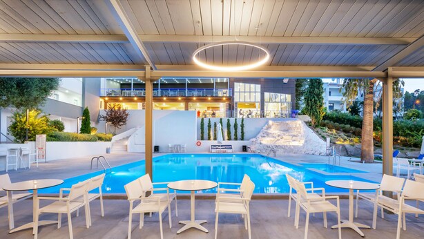 Un área de piscina con sillas y mesas blancas, un edificio moderno al fondo y un cielo despejado.