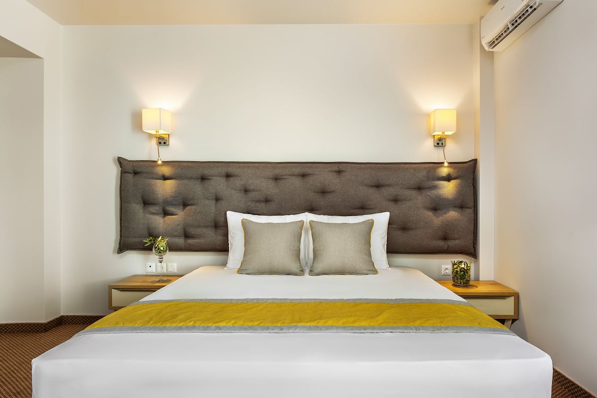 Ein Hotelzimmer mit einem großen Bett, zwei Nachttischlampen, einem kleinen Tisch mit einer Vase und einer Klimaanlage an der Wand.