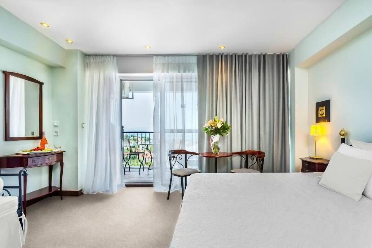 Habitación de hotel con cama, escritorio, silla, mesita con un jarrón, mesita de noche con lámpara y balcón con cortinas.