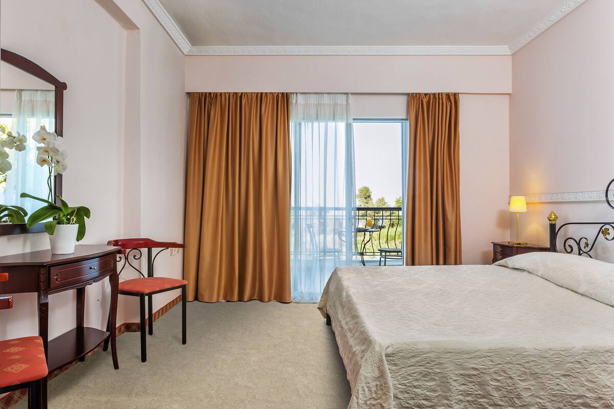 Ein Hotelzimmer mit Bett, Schreibtisch mit Stuhl, einer Zimmerpflanze und einem Balkon mit Vorhängen.