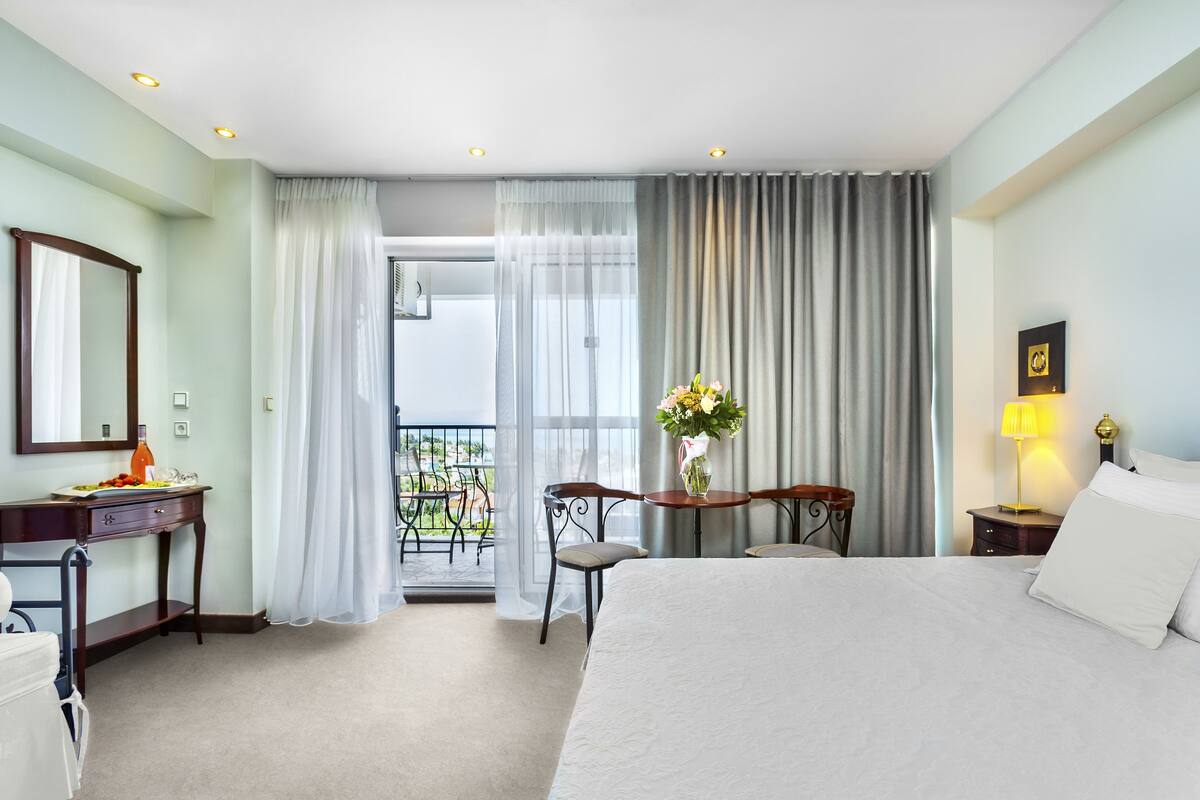 Ein Hotelzimmer mit einem Bett, einem Schreibtisch, einem Sessel, einem Tisch mit Blumen und einem Balkon mit Blick.