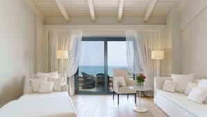 A hotel room with a large sliding glass door leading to a balcony with a view of the sea. There are two white sofas, a small round table with a vase of flowers, and two floor lamps. - Aegean Pearl (Rethymno)